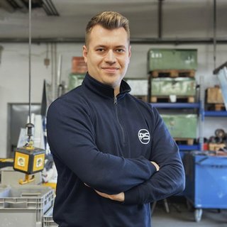 Ein junger Mann mit kurzem, blondem Haar steht in einem industriellen Umfeld und lächelt selbstbewusst in die Kamera.
