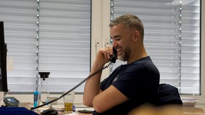 In einem modernen Büro telefoniert ein lächelnder Mann, während er an seinem Schreibtisch sitzt und auf den Bildschirm schaut.