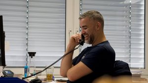 In einem modernen Büro telefoniert ein lächelnder Mann, während er an seinem Schreibtisch sitzt und auf den Bildschirm schaut.