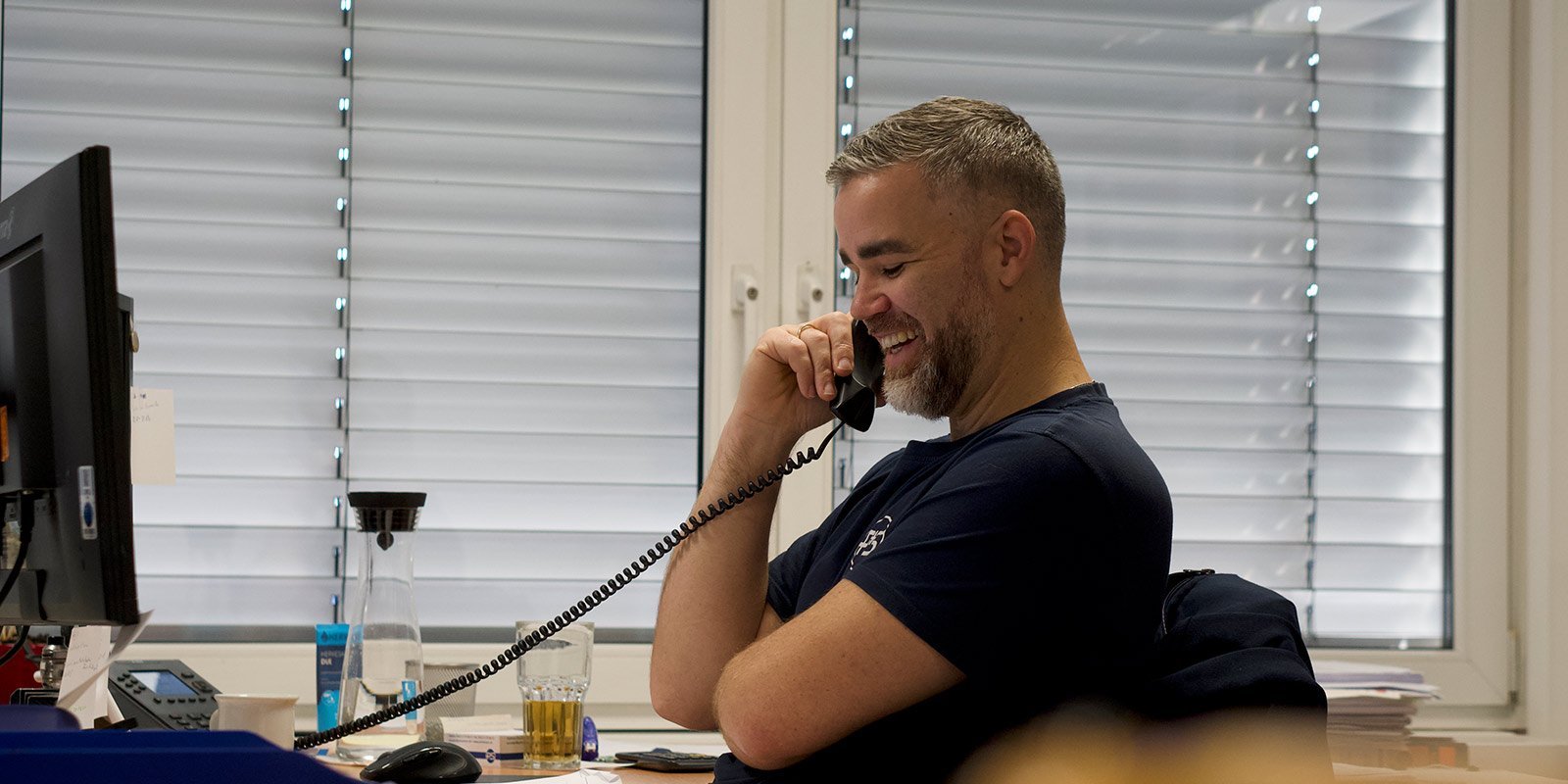 In einem modernen Büro telefoniert ein lächelnder Mann, während er an seinem Schreibtisch sitzt und auf den Bildschirm schaut.