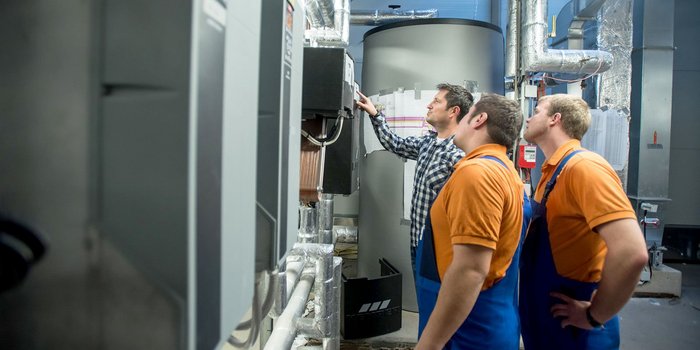 Drei Techniker in blauen Overalls und orangefarbenen T-Shirts betrachten aufmerksam eine technische Anlage in einem modernen Raum.