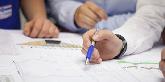 Mehrere Hände sind um einen Tisch versammelt, während eine Person einen Plan mit einem blauen Stift bearbeitet.