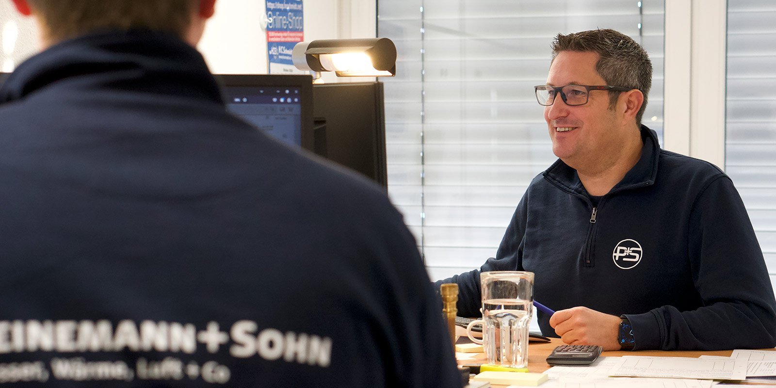 Ein lächelnder Mann sitzt an einem Schreibtisch, während er mit einem Kollegen spricht, der ihm den Rücken zuwendet.