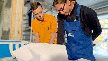 Zwei Männer, einer in einem orangefarbenen Shirt und der andere in blauer Arbeitskleidung, studieren gemeinsam Dokumente.