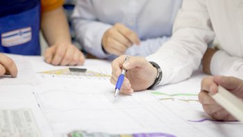 Mehrere Hände sind über technische Pläne gebeugt, während eine Person mit einem blauen Stift Notizen macht.