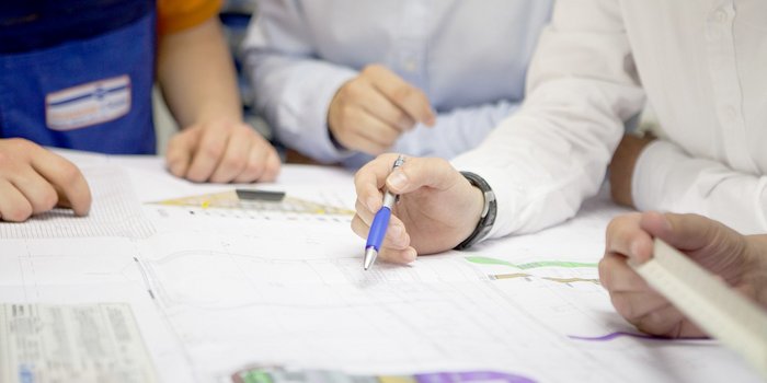 Mehrere Hände sind über technische Pläne gebeugt, während eine Person mit einem blauen Stift Notizen macht.