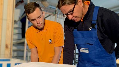 Ein junger Mann in einem orangefarbenen Shirt und ein älterer Mann in blauer Arbeitskleidung betrachten aufmerksam einen Plan.
