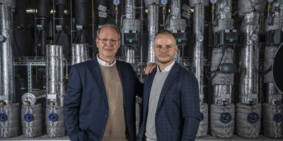 Zwei Männer stehen in einem industriellen Umfeld, umgeben von isolierten Rohren und Messgeräten, die eine technische Atmosphäre schaffen.