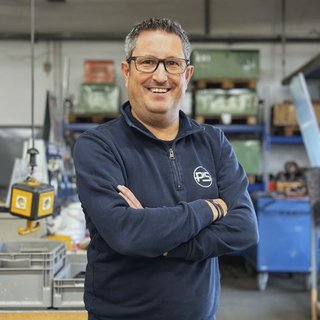 Ein lächelnder Mann mit Brille steht in einer Werkstatt, umgeben von Werkzeugen und Kisten, und hat die Arme verschränkt.