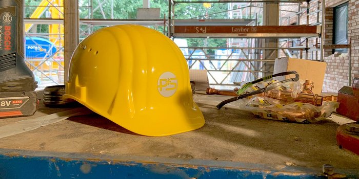 Auf einem Baustelltisch sind ein gelber Helm, eine Bohrmaschine und verschiedene Kupferteile sichtbar, die auf Arbeit hinweisen.