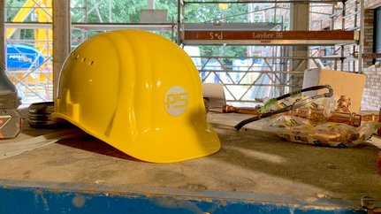 Auf einem Baustelltisch sind ein gelber Helm, eine Bohrmaschine und verschiedene Kupferteile sichtbar, die auf Arbeit hinweisen.