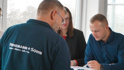 Ein Mann mit Brille und ein weiterer Mann besprechen Dokumente an einem Tisch, während eine Frau im Hintergrund zuschaut.