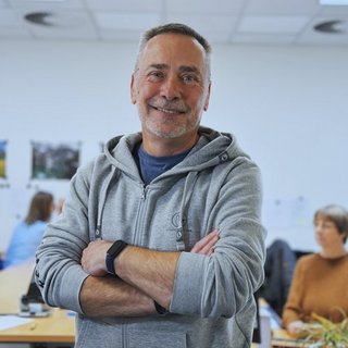 Ein lächelnder Mann in einem grauen Hoodie steht mit verschränkten Armen in einem hellen Büro voller Menschen.