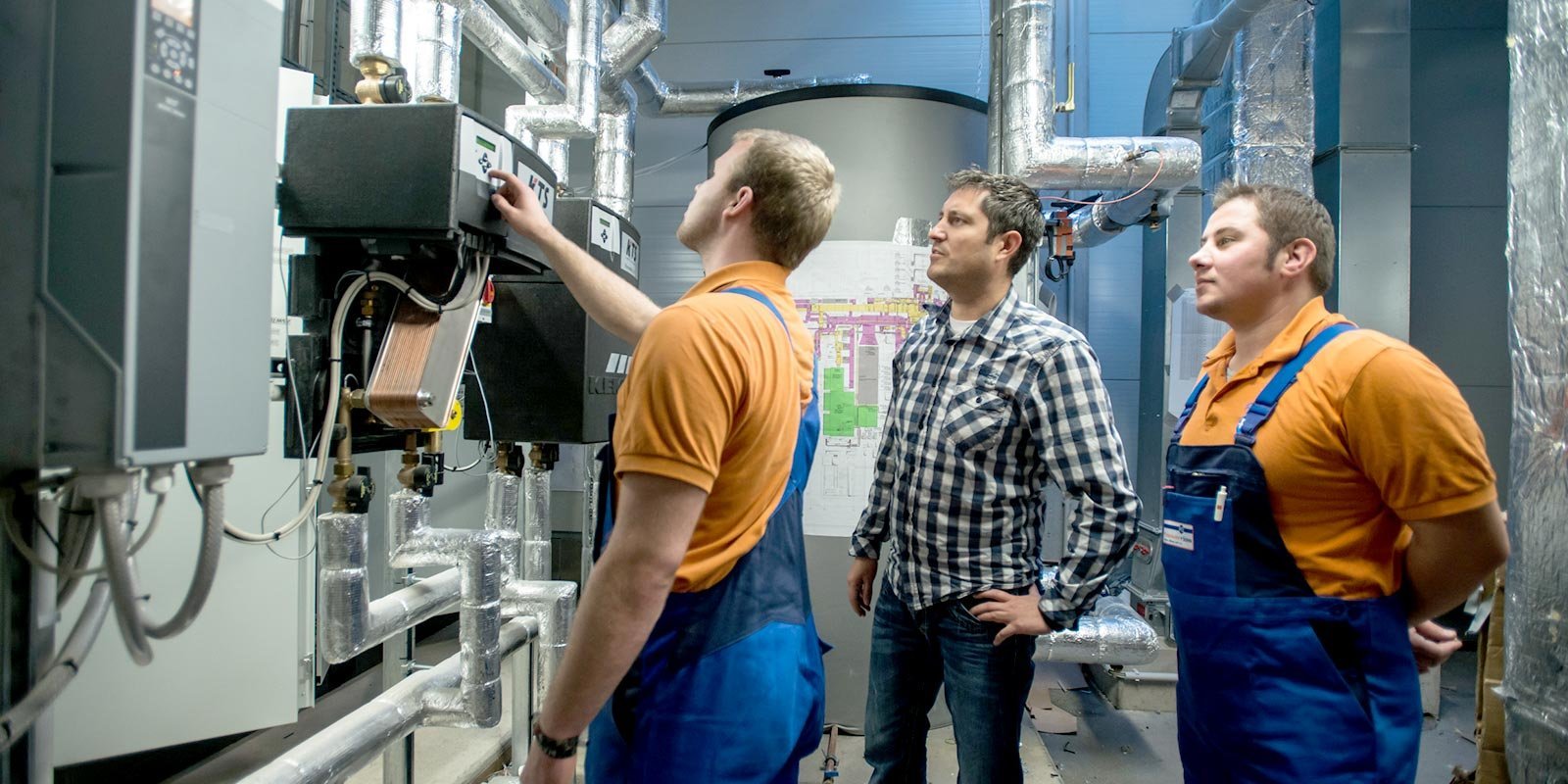 Ein Techniker justiert ein Gerät, während zwei weitere Männer aufmerksam zuschauen, umgeben von Rohren und Maschinen.
