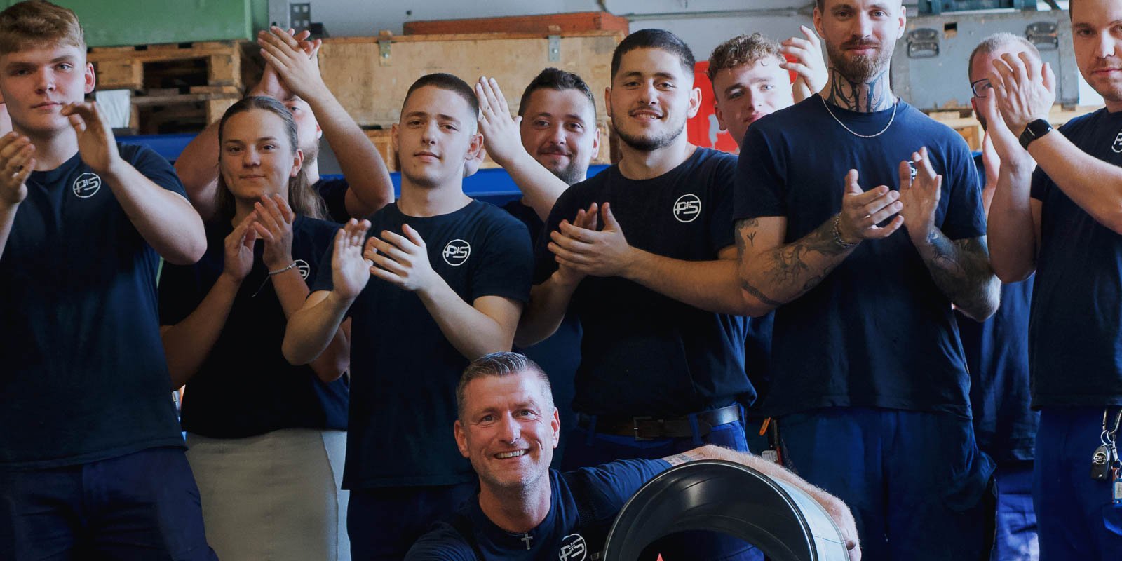 Eine Gruppe von jungen Menschen in blauen T-Shirts applaudiert begeistert, während ein Mann lächelnd im Vordergrund sitzt.