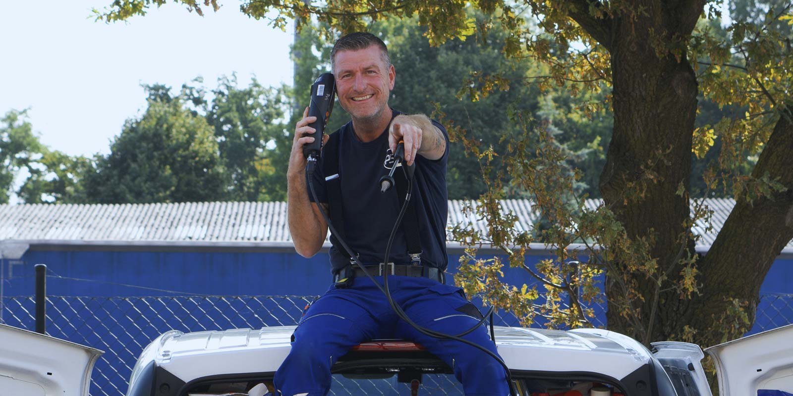 Ein fröhlicher Mann sitzt auf dem Dach eines Autos und hält ein Werkzeug in der Hand, während er in die Kamera lächelt.