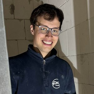 Ein junger Mann mit Brille lächelt fröhlich, während er zwischen Metallrahmen in einem Baustellenumfeld steht.