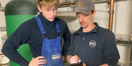 Zwei Männer in Arbeitskleidung betrachten gemeinsam ein Tablet in einem technischen Raum mit Rohren und Geräten im Hintergrund.