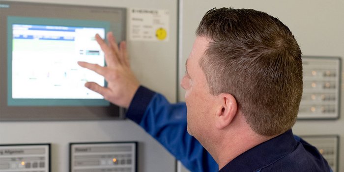 Ein Mann in blauer Arbeitskleidung bedient einen digitalen Bildschirm, während er auf die Anzeige zeigt.