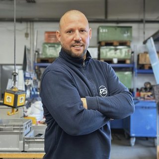 Der Mann in blauer Arbeitskleidung posiert selbstbewusst mit verschränkten Armen in einer gut ausgestatteten Werkstatt.