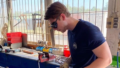 Der Mann trägt eine Schutzbrille und zeigt eine ernste Miene, während er an einem Projekt in einer gut beleuchteten Werkstatt arbeitet.