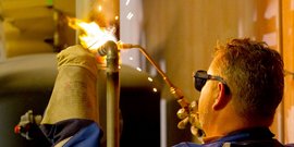 Ein Handwerker schweißt mit einer Flamme, während Funken in der Luft sprühen, umgeben von industrieller Umgebung.