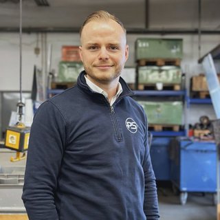 In einer industriellen Umgebung lächelt ein junger Mann in einem blauen Pullover, während er vor Regalen mit Werkzeugen posiert.