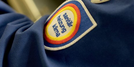 Ein blauer Stoff mit einem aufgenähten Emblem, das die Worte 'Sanitär Heizung Klima' zeigt, ist gut sichtbar.