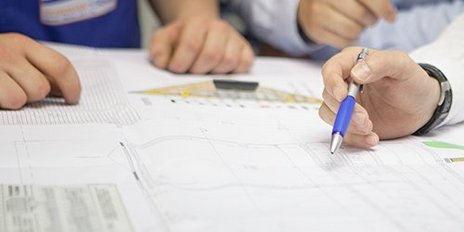 Auf einem Tisch liegen Baupläne, während zwei Personen intensiv über die Details der Zeichnungen sprechen.