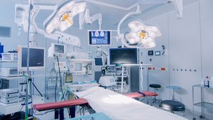 Ein moderner Operationssaal mit mehreren Monitoren, chirurgischen Lampen und einem vorbereiteten OP-Tisch, der mit einem weißen Tuch bedeckt ist.