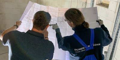 Zwei Personen studieren gemeinsam einen Bauplan in einem hellen Raum mit weißen Wänden und einem Fenster im Hintergrund.
