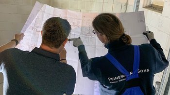 Zwei Personen studieren gemeinsam einen Bauplan in einem hellen Raum mit weißen Wänden und einem Fenster im Hintergrund.