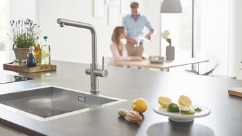 Der Wasserhahn in der Küche steht im Fokus, während eine Frau und ein Mann entspannt am Tisch sitzen und sich unterhalten.