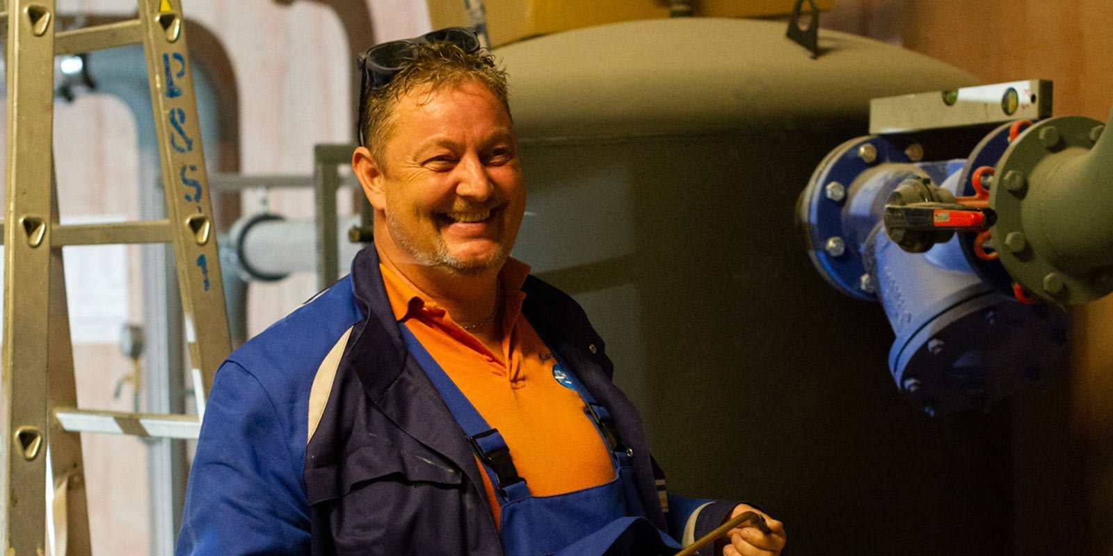 Ein lächelnder Mann in Arbeitskleidung steht in einer industriellen Umgebung, umgeben von Rohren und Maschinen.