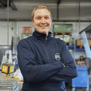In einer industriellen Umgebung lächelt ein Mann selbstbewusst, während er in einem blauen Pullover gekleidet ist.