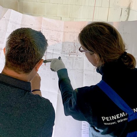 Zwei Personen studieren gemeinsam einen Bauplan, während sie in einem hellen Raum mit weißen Wänden stehen.