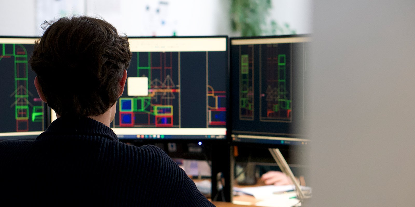 In einem modernen Büro arbeitet ein Mann an Computerbildschirmen, die komplexe Entwürfe und Pläne darstellen.
