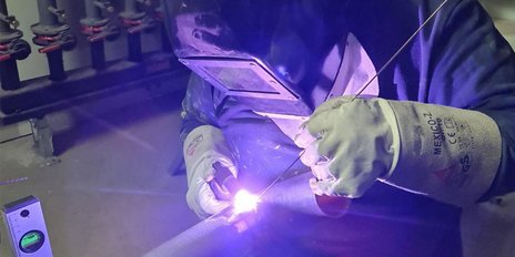 In einer Werkstatt schweißt eine Person mit Schutzhandschuhen und einem Helm, während violettes Licht den Arbeitsbereich erhellt.