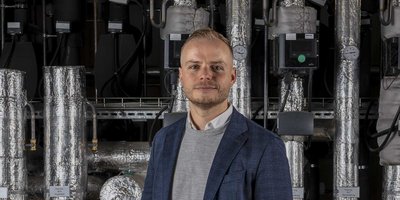 Ein Mann in einem blauen Blazer steht vor einer komplexen Heizungsanlage mit isolierten Rohren und Messgeräten.