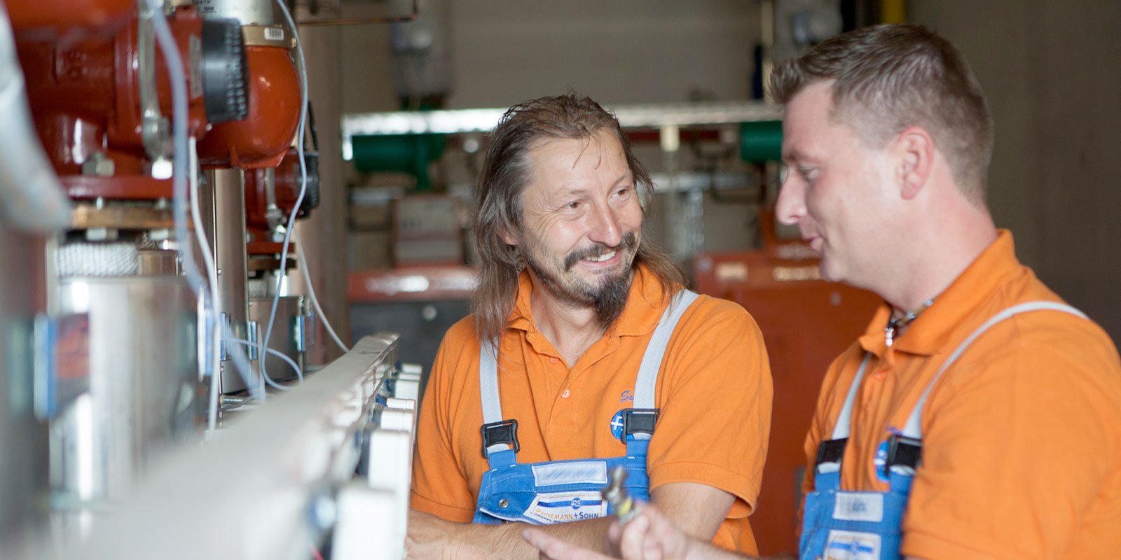 Zwei Männer in orangefarbenen Shirts und blauen Latzhosen stehen an einer Maschine und unterhalten sich freundlich.