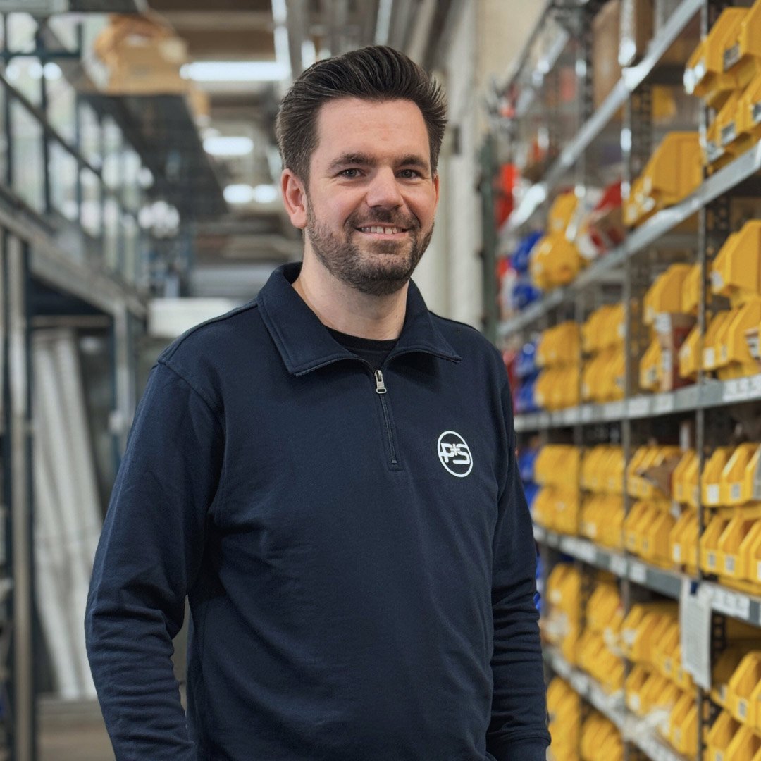 Ein Mann mit Bart lächelt in einem Lager, umgeben von Regalen voller gelber und blauer Behälter.