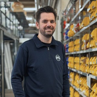 Ein Mann mit Bart lächelt in einem Lager, umgeben von Regalen voller gelber und blauer Behälter.