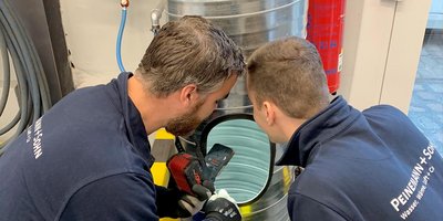 Zwei Männer in Arbeitskleidung blicken konzentriert in einen großen Metallbehälter, während einer ein Werkzeug hält.