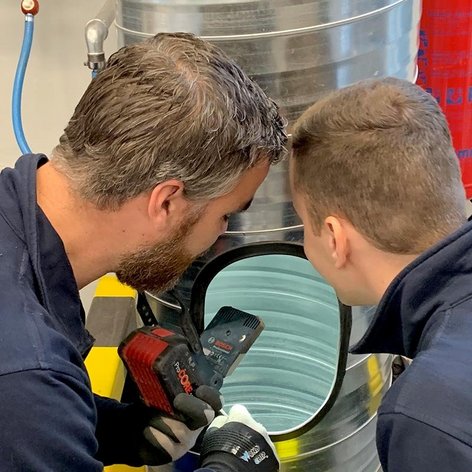 Zwei Männer in Arbeitskleidung blicken konzentriert in einen großen Metallbehälter, während einer ein Werkzeug hält.