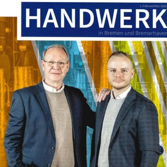 Zwei Männer stehen nebeneinander, lächelnd, in formeller Kleidung, vor einem bunten, industriellen Hintergrund.