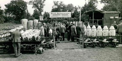 Eine Gruppe von Menschen betrachtet landwirtschaftliche Geräte auf einer Ausstellung, umgeben von Bäumen und Grünflächen.