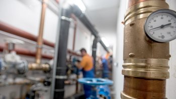 Im Vordergrund ist ein Messgerät zu sehen, während Techniker in blauen Overalls an der Rohrleitung arbeiten.