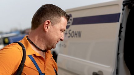 Ein Mann mit einem orangefarbenen Shirt und einer Umhängetasche steht neben einem Lieferwagen und schaut nachdenklich.