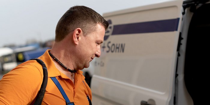 Ein Mann mit einem orangefarbenen Shirt und einer Umhängetasche steht neben einem Lieferwagen und schaut nachdenklich.