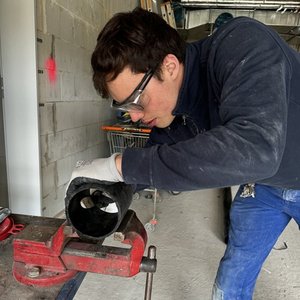 Ein junger Mann arbeitet konzentriert an einem Rohr, das in einem Schraubstock fixiert ist, während er Schutzhandschuhe trägt.